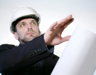 Male with white hard hat, dark suit, four o'clock shadow and hand outstretched toward set of plans.