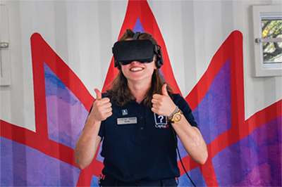 A young woman is wearing a virtual reality headset. She is smiling and giving two thumbs up.