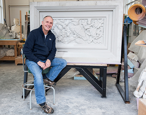 A man in a workshop sits in front of a large stone carving that features an image of an owl.