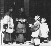 Photographie de quatre enfants portant le costume traditionnel chinois, debout devant une porte de magasin en brique