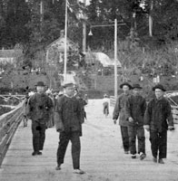 Photographie de cinq hommes chinois qui se promnent sur un trottoir en bordure de l'eau, fort en arrire-plan, Vancouver (Colombie-Britannique), 24 mai 1899