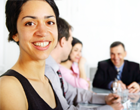 Smiling young woman in foreground and a man facing smiling at head of table and contemplative second woman.