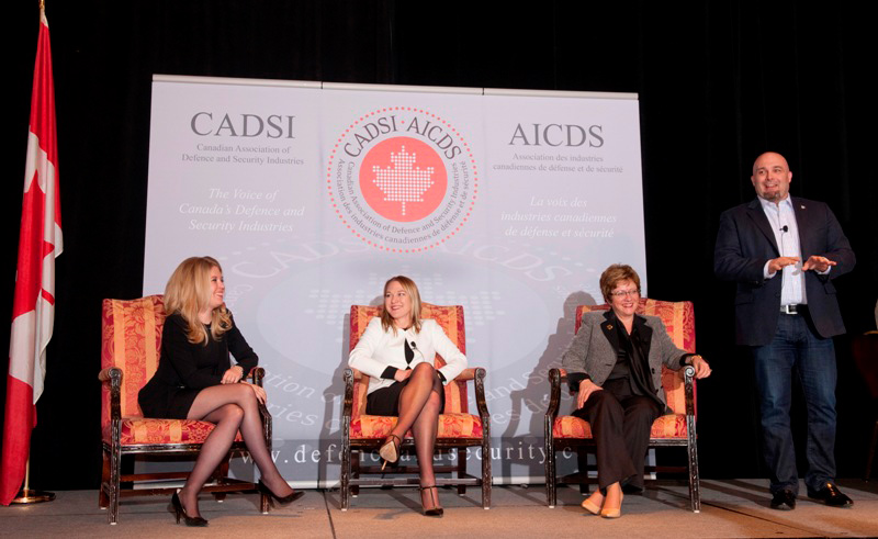 Three women sitting on a stage while a man stands and talks.