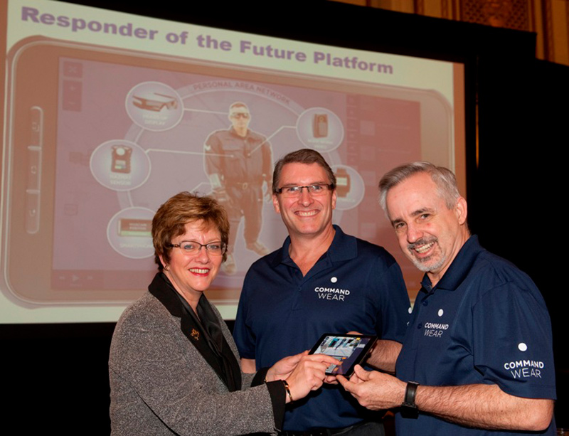 A woman and two men holding a tablet while smiling at the camera.