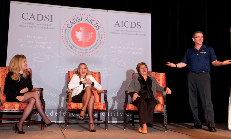 Three women sitting and laughing on a stage while a man stands and smiles.