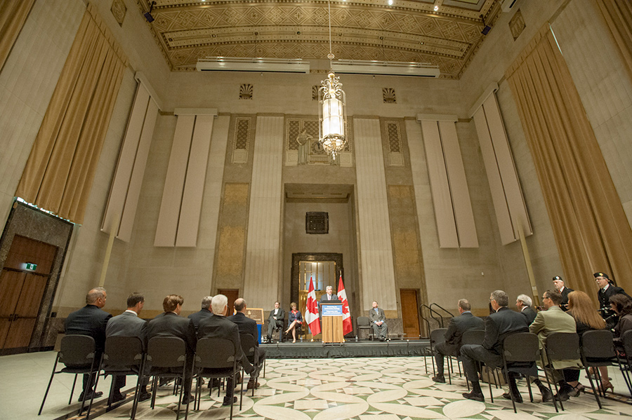 Man standing behind a podium, a woman and two men sitting on stage while a crowd looks at them.