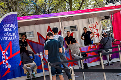 Four people are sitting at the virtual reality kiosk outside on the plaza of 90 Wellington. The kiosk consists of four stations along the outside of a painted shipping container.