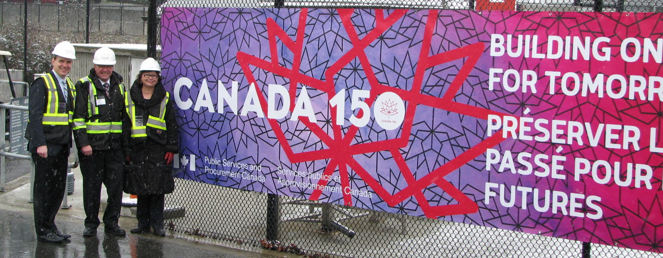 Deux hommes et une femme se tiennent debout et sourient à côté d’une bannière de Canada 150.