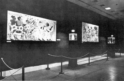 The first gallery, south wall. In the first two rooms direct lighting was limited to the objects themselves leaving the rest of the galleries in semi-darkness. This method greatly dramatized the objects and emphasized their individual beauty. Photographic reproductions of tomb frescoes and enlargements of the details of objects formed a background to the widely spaced display cases.